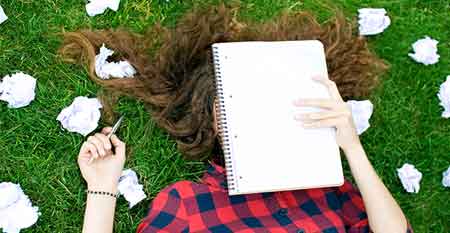 A woman lying on the grass doing homework, crumpled paper around her
